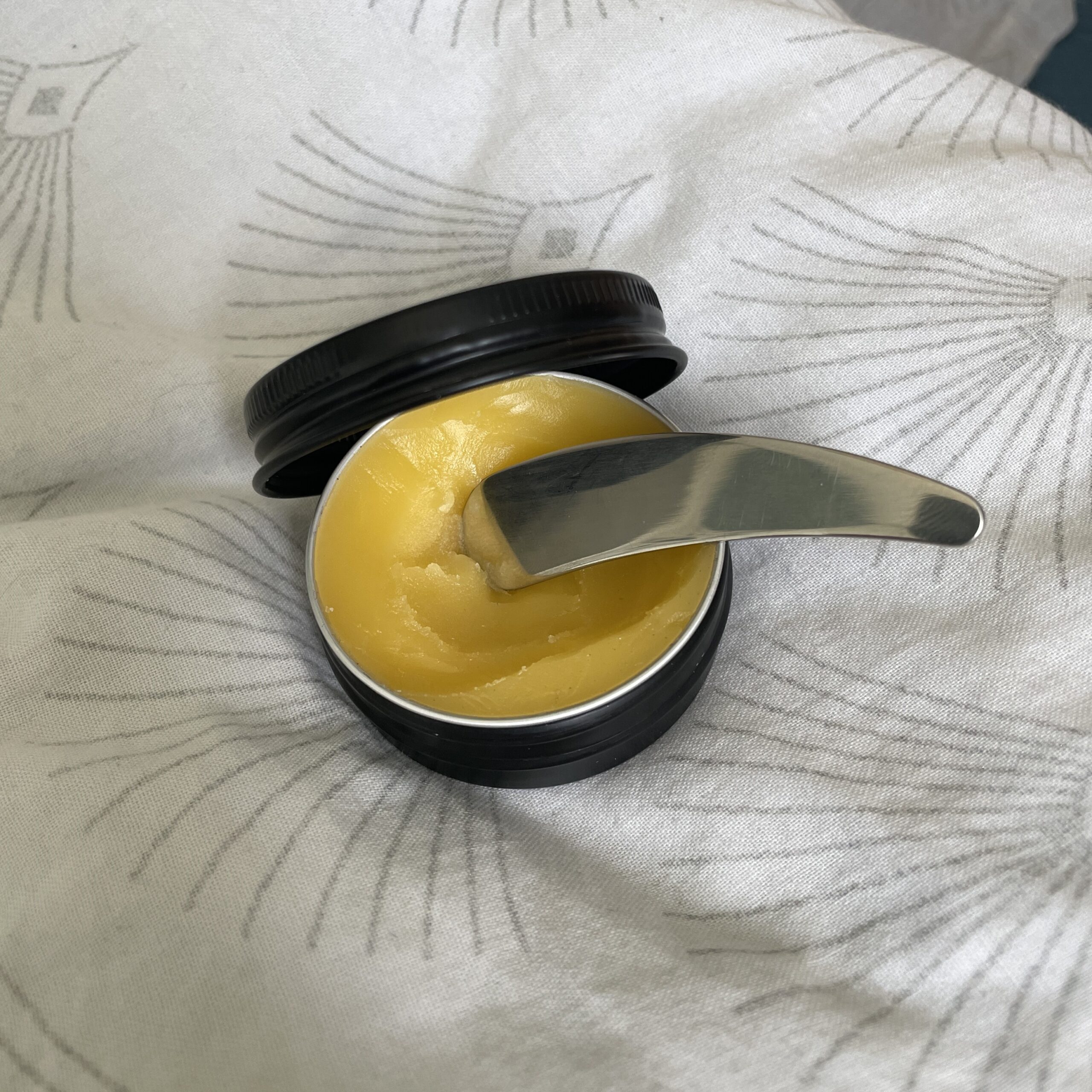 Pot rond noir ouvert laissant apparaitre un baume corporel jaune, une spatule en inox dedans